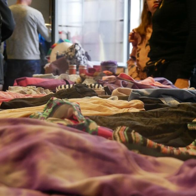 Tisch mit ausgebreiteten Kleidungsstücken auf einem Flohmarkt, im Hintergrund stehen mehrere Personen.
