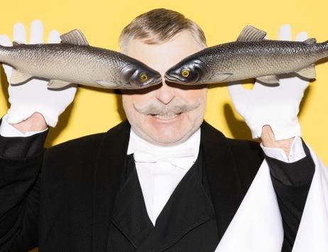 Ein Mann in einem weißen Hemd, weißen Handschuhen und einem schwarzen Jacket steht vor einem gelben Hintergrund. Er hält in jeder Hand einen Fisch, dessen Köpfe vor seinen eigenen Augen liegen.