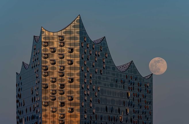 Das Foto zeigt die Elbphilharmonie in Hamburg im Abendlicht, auf deren äußerster Dachzacke ein Vollmond balanciert.