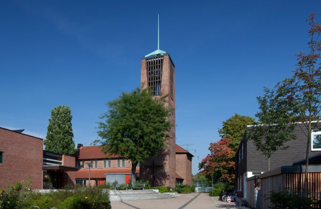 Eine Kirche mit einem hohen Turm und rotem Ziegeldach, umgeben von Bäumen und einem gepflasterten Platz.