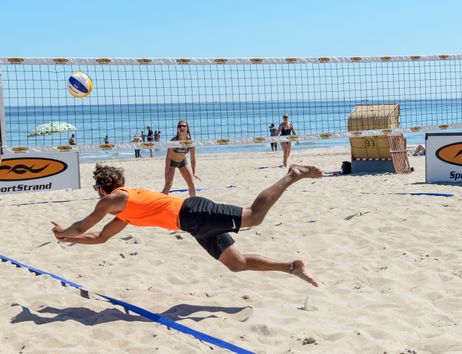 Ein Mann in einem orangefarbenen Shirt springt nach einem Volleyball am Strand. Im Hintergrund sind ein Netz, Strandkörbe und das Meer zu sehen.