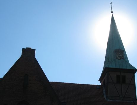 Kirchturm mit Uhr vor strahlend blauem Himmel und Sonne im Hintergrund.