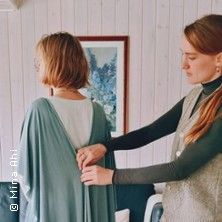 Zwei Frauen in einem Raum, eine hilft der anderen beim Anziehen eines blauen Kleides.