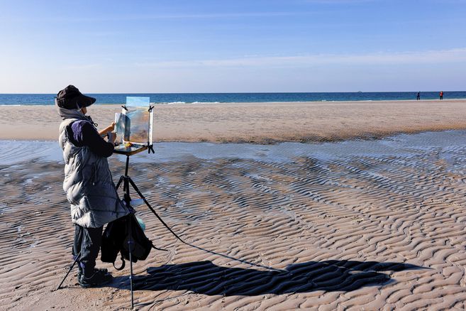 Eine Frau steht mit einer Staffelei am Strand und malt auf eine Leinwand.