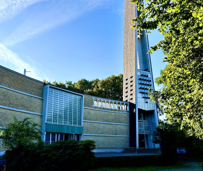 Außenansicht der Dreifaltigkeitskirche mit modernem Turm und Glasfassade, umgeben von Bäumen.