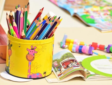 Ein gelber Behälter mit bunten Buntstiften, ein offenes Kinderbuch und bunte Perlen auf einem Tisch.