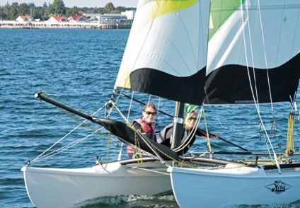 Zwei Personen segeln auf einem Katamaran auf einem See mit Küste im Hintergrund.