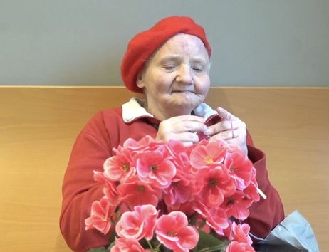 Ältere Frau in roter Kleidung und rotem Barett hält einen großen Strauß pinker Blumen und arbeitet mit den Händen daran.
