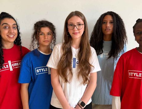 Five people with different hair lengths and textures are wearing colorful T-shirts with the words “STYLE CREATE” printed on them and are standing in front of a light-colored wall.