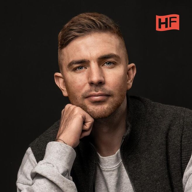 Porträt eines Mannes mit kurzem Haar und Bart, der nachdenklich in die Kamera schaut. Er trägt einen grauen Pullover und eine dunkle Weste. Im Hintergrund ist ein HF-Logo.