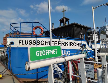 Grünes Schild mit weißer Aufschrift "Geöffnet" vor dem blau-weißen Profil der Flussschifferkirche im Hamburger Hafen.
