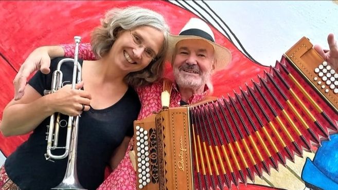 Ein Mann mit Akkordeon und eine Frau mit Trompete lächeln in die Kamera vor einem bunten Hintergrund.