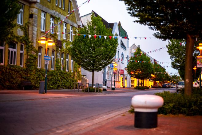 Eine ruhige, mit Bäumen gesäumte Straße in einer Stadt bei Dämmerung, geschmückt mit bunten Wimpeln und beleuchtet von Straßenlaternen.