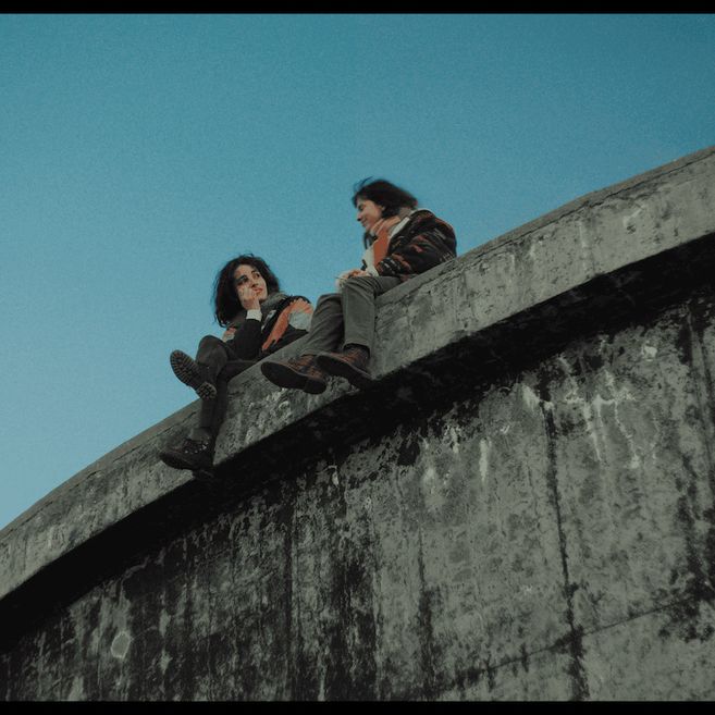 Zwei Personen sitzen auf dem Rand einer großen Betonstruktur vor blauem Himmel, passend zum Titel „37. Hamburg International Queer Film Festival - Miss Carbón (Queen of Coal) | Eröffnungsfilm“.