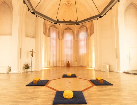 Innenansicht eines hellen Kirchenraums mit Meditationskissen und Bänken auf dem Boden.