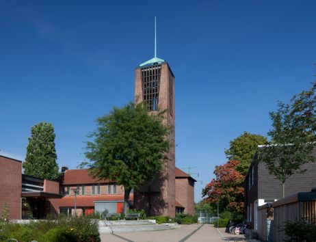 Eine Kirche mit einem hohen Turm und rotem Ziegeldach, umgeben von Bäumen und einem gepflasterten Platz.