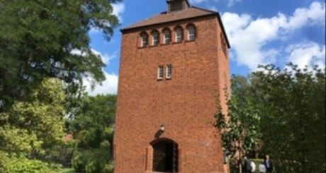 Ein roter Backsteinturm mit einem gewölbten Eingang und mehreren Fenstern, umgeben von Bäumen und einem blauen Himmel im Hintergrund.