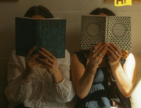 Zwei Personen sitzen auf einem Sofa und lesen Bücher, deren Cover ihre Gesichter verdecken. Oben rechts ist ein gelbes Logo mit den Buchstaben 'HF'.