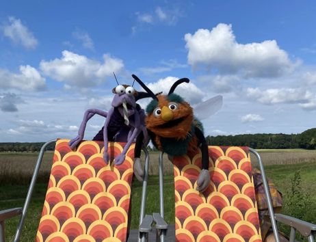 Zwei bunte Puppen, eine lila und eine orange, sitzen auf Liegestühlen in einer Wiese unter blauem Himmel.