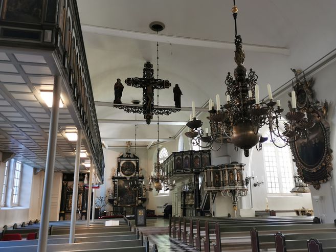 Innenansicht einer Kirche mit Emporen, Kronleuchtern, Kreuz und Altarraum; leere Bänke im Vordergrund.