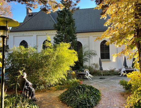Ein ruhiger Garten mit einer weißen Kirche im Hintergrund, umgeben von Bäumen und Pflanzen. Eine Person sitzt auf einer Bank im Vordergrund.