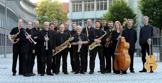 Eine Jazzband posiert mit ihren Instrumenten im Freien vor einem modernen Gebäude.