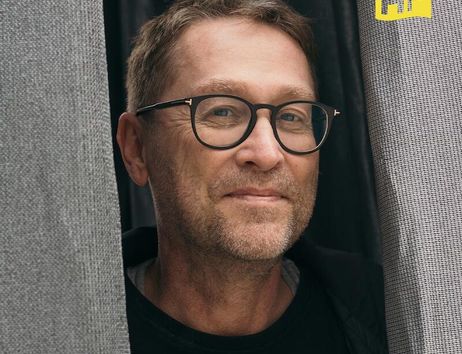 Ein Mann mit Brille lächelt zwischen zwei grauen Vorhängen hervor. Oben rechts ist ein gelbes Logo mit den Buchstaben 'HF' zu sehen.