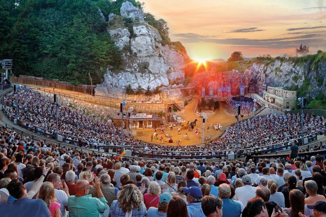 A large crowd is seated in an outdoor theater, surrounded by rocky landscapes. The sun is setting in the background, casting a warm light over the scene.
