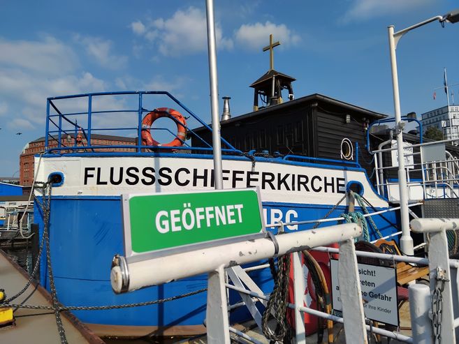 Grünes Schild mit weißer Aufschrift "Geöffnet" vor dem blau-weißen Profil der Flussschifferkirche im Hamburger Hafen.