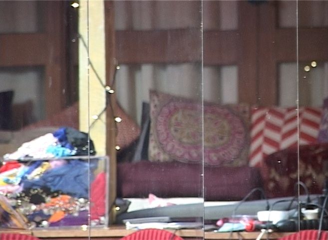 Ein Film Still zeigt die Reflektion von verschieden gemusterten Kissen, die im Hintergrund vor einer Hausfassade aufgetürmt sind. Im Vordergrund lässt sich eine Tischplatte erahnen, auf der viele Kabel, Papiere, eine durchsichtige Kiste mit Textilien und weiterer Kram liegt.
