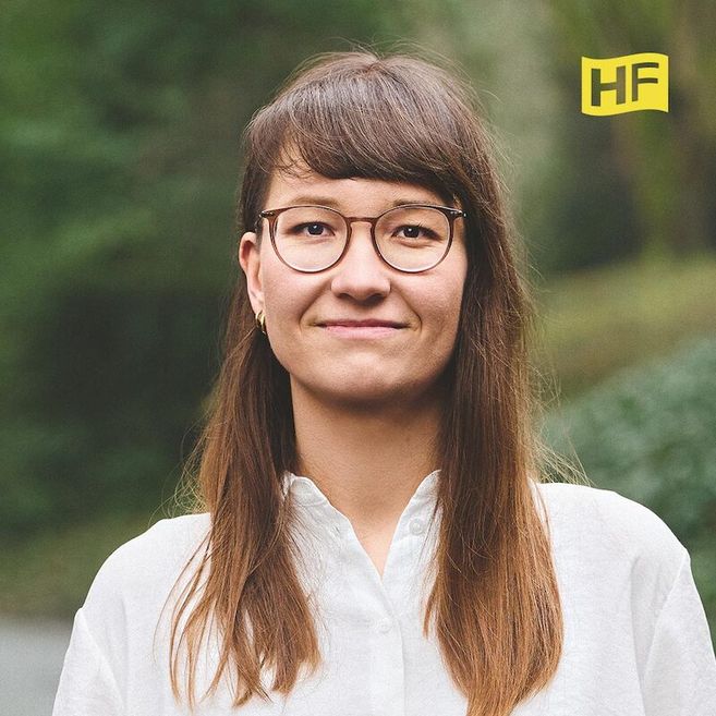 Porträt einer Frau mit Brille und langen Haaren, die ein weißes Hemd trägt. Im Hintergrund ist verschwommenes Grün zu sehen. Oben rechts ist ein gelbes Logo mit den Buchstaben 'HF'.