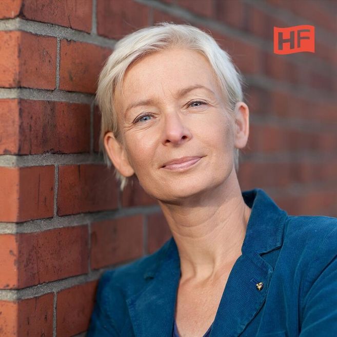 Porträt einer Frau mit kurzem blondem Haar, die an eine Backsteinmauer lehnt. Sie trägt einen blauen Blazer. Oben rechts ist ein rotes Logo mit den Buchstaben 'HF'.