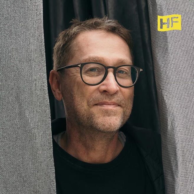 Ein Mann mit Brille lächelt zwischen zwei grauen Vorhängen hervor. Oben rechts ist ein gelbes Logo mit den Buchstaben 'HF' zu sehen.