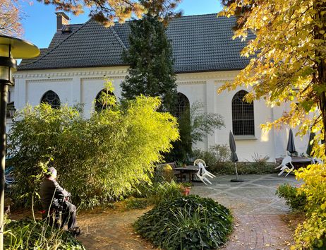 Ein weißes Gebäude mit großen Fenstern, umgeben von herbstlichen Bäumen und einem kleinen Garten mit Sitzgelegenheiten.
