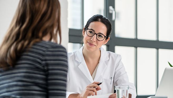 Es sind zwei Frauen zu sehen, eine sitzt vorne im Bild und ist von hinten zu sehen und eine sitzt in einem Hemd oder Arztkittel der anderen Frau gegenüber.