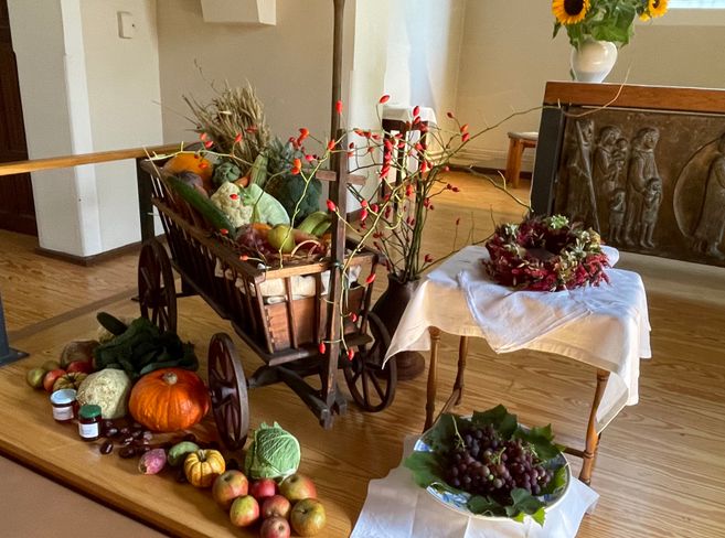 Ein Erntealtar mit einem Holzwagen voller Gemüse und Obst, umgeben von herbstlichen Dekorationen und einem Tisch mit Trauben und einem Kranz.