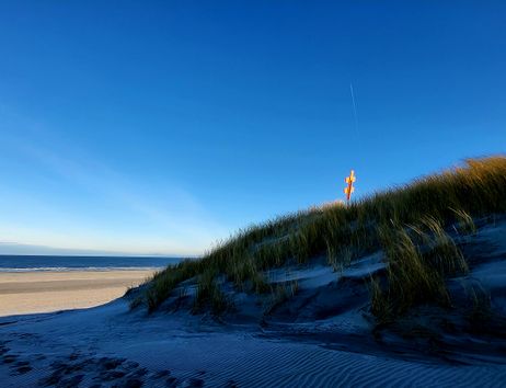 Dünenlandschaft am Strand mit Meer und blauem Himmel; auf dem Hügel steht ein rot-weißes Seezeichen.