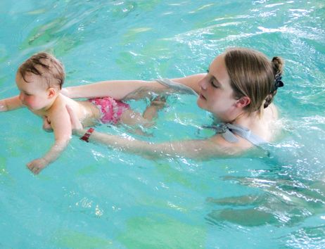 Eine Frau hält ein Baby im Wasser, während es nach einem gelben Ball greift.
