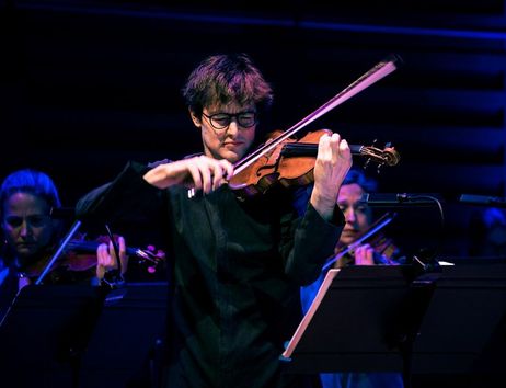 Ein Geiger spielt konzentriert auf der Bühne, umgeben von anderen Musikern im Hintergrund.