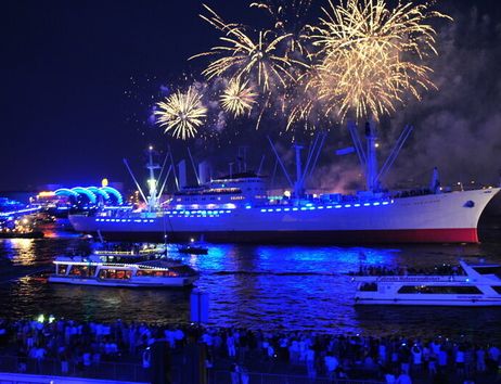 Ein beleuchtetes Schiff mit Feuerwerk am Nachthimmel in einem Hafen.