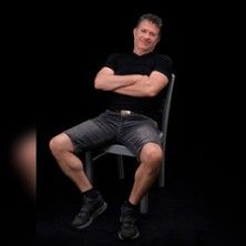 Ein Mann sitzt lässig auf einem Stuhl mit verschränkten Armen und Beinen, vor einem schwarzen Hintergrund.