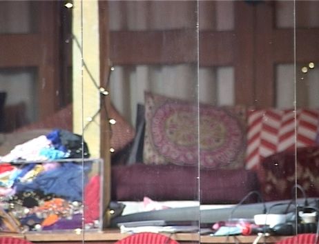 Ein Film Still zeigt die Reflektion von verschieden gemusterten Kissen, die im Hintergrund vor einer Hausfassade aufgetürmt sind. Im Vordergrund lässt sich eine Tischplatte erahnen, auf der viele Kabel, Papiere, eine durchsichtige Kiste mit Textilien und weiterer Kram liegt.