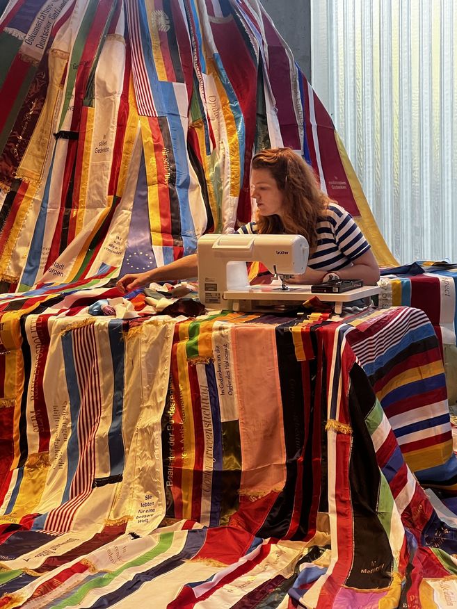 Eine Frau näht an einer Nähmaschine an einem großen, bunten Vorhang aus vielen bedruckten Stoffbändern mit Erinnerungs- und Gedenktexten.