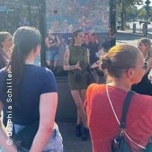 Eine Gruppe von Menschen steht im Freien und hört einer Frau zu, die spricht. Im Hintergrund ist eine Karte oder ein Plakat zu sehen.