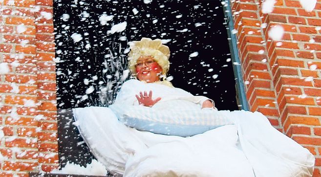 Eine Person in einem Frau Holle Kostüm schüttelt Kissen aus einem Fenster eines Backsteingebäudes, wodurch es schneit.