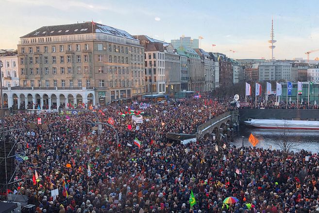 Große Menschenmenge bei einer Demonstration in einer Innenstadt am Wasser, mit vielen Fahnen und Transparenten; im Hintergrund ist der Fernsehturm Hamburg zu sehen.