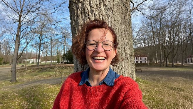 Eine lächelnde Person mit Brille und rotem Pullover steht vor einem Baum in einem Park.