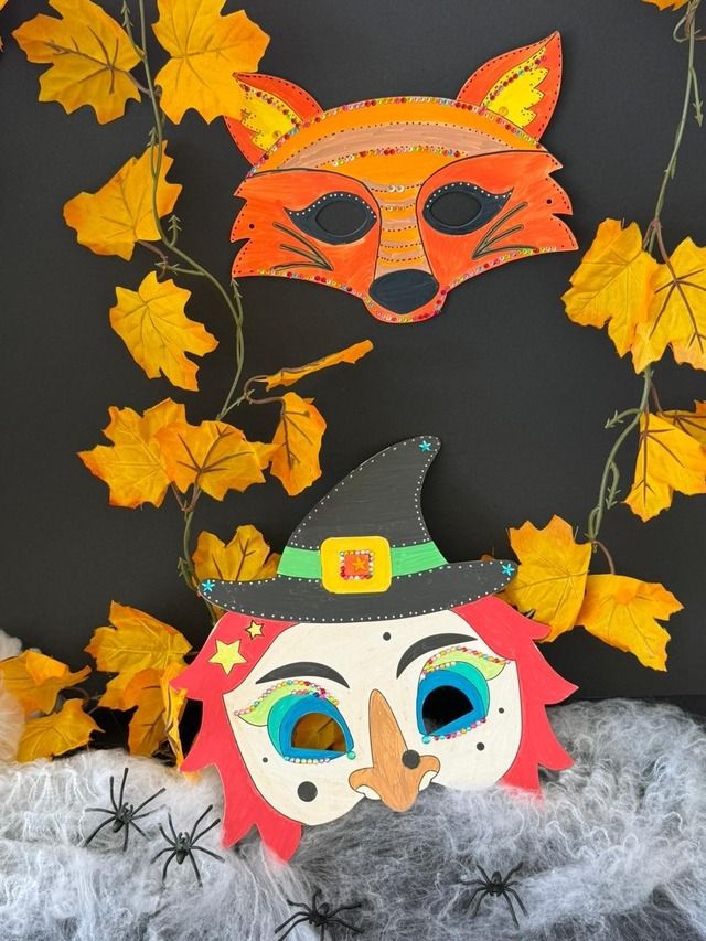 Zwei bunte Masken, eine Fuchsmaske und eine Hexenmaske, hängen an einer Wand mit Herbstblättern und Spinnweben.