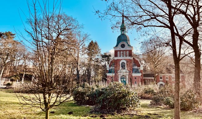 Eine rote Backsteinkirche mit grünem Turm, umgeben von Bäumen und Sträuchern im Frühling.