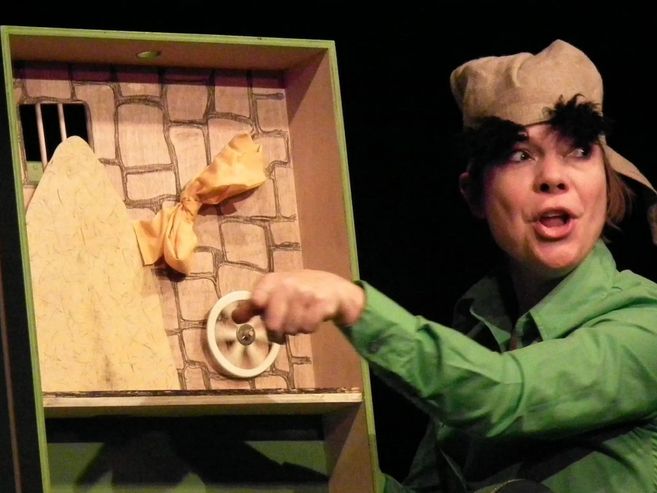Eine Person in einem grünen Kostüm spielt in einem Puppentheater. Sie trägt eine Mütze und zeigt auf ein hölzernes Bühnenbild mit einem Rad.